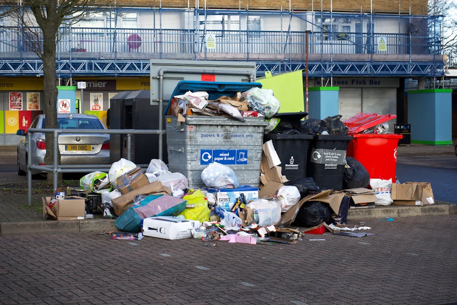 A collection of overflowing rubbish bins and scattered waste surrounding a large grey mixed paper and card recycling container situated on a paved urban street. The recycling bin is partially open, re