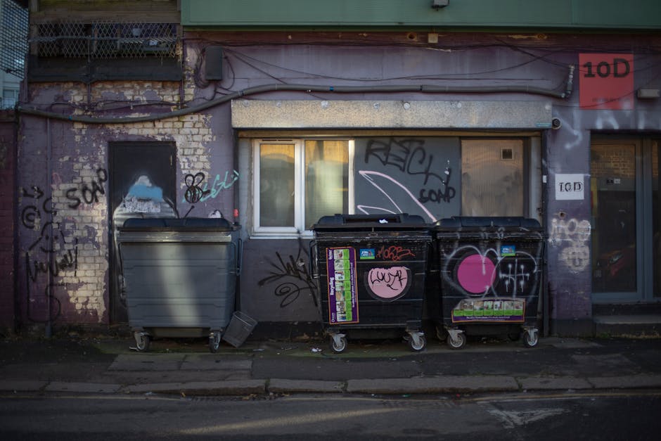 The image depicts a section of an urban building facade featuring a small, rectangular window with a white frame, situated between two graffiti-covered walls. The wall on the left shows exposed brickw