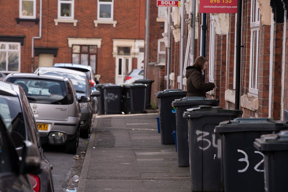 The image shows the rear section of a white commercial van parked on a city street, with three large blue plastic rubbish bags placed on the ground next to it. The bags are filled with waste material